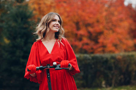 Young woman staying with electric scooter in red dress and smile at the autumn city parkの写真素材