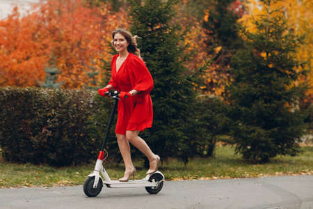 Young woman with electric scooter in red dress at the autumn city parkの写真素材