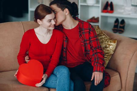 Smiling young woman and man with valentines gift box red heart shaped at home sofaの写真素材