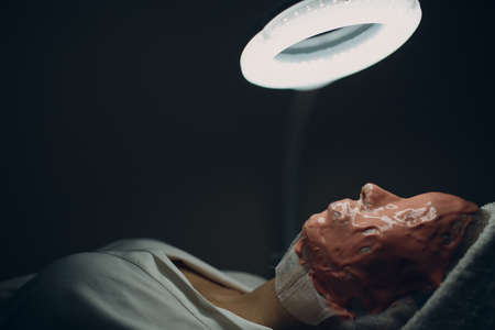 Alginate face mask application process. A female hands of a beautician holding spatula. Woman lying on a couch in office of cosmetologist. Facial skincare in the beauty spa salonの写真素材