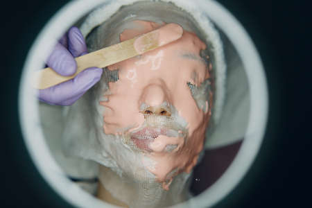 Alginate face mask application process. A female hands of a beautician holding spatula. Woman lying...