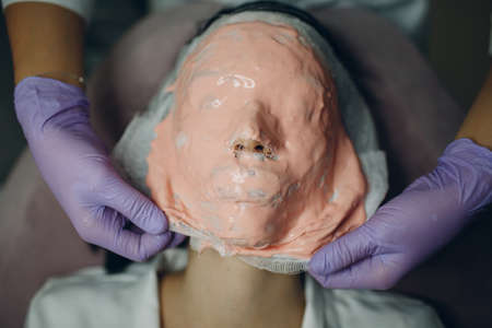 Alginate face mask removal process. A female hands of a beautician holding spatula. Woman lying on a couch in office of cosmetologist. Facial skincare in the beauty spa salonの写真素材