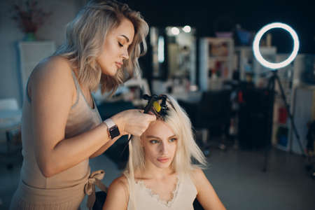 Hairdresser female making hair extensions to young woman with blonde hair in beauty salon. Professional hair extension.の写真素材