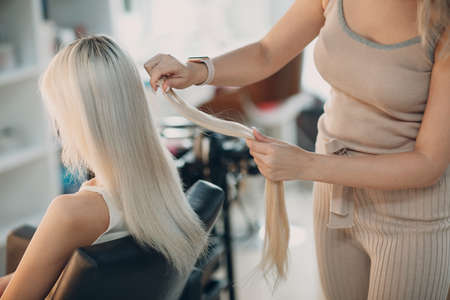 Hairdresser female making hair extensions to young woman with blonde hair in beauty salon. Professional hair extension.の写真素材