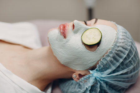 Young woman applying mask of clay on face with cucumber on eyes in beauty spaの写真素材