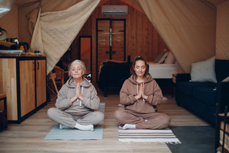 Woman senior and young relaxing at glamping camping tent. Women family elderly mother and young daughter doing yoga and meditation indoor. Modern zen-like vacation lifestyle concept.の写真素材