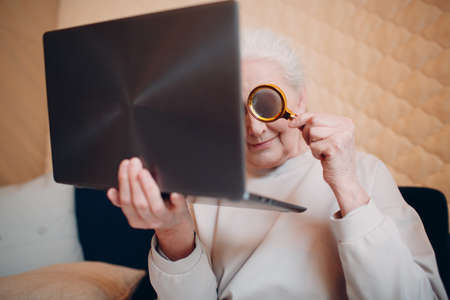 Elderly woman grandmother learns to work at home on laptop computer and internet with magnifierの写真素材