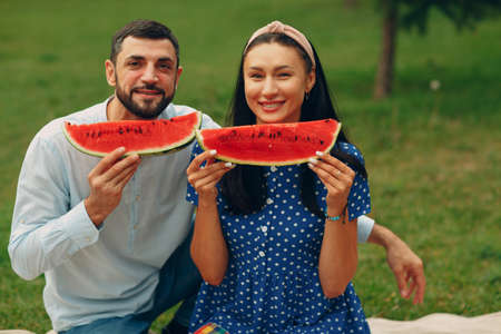 Young adult woman and man couple picnic at green grass meadow in park having fun and smile with watermelonの写真素材