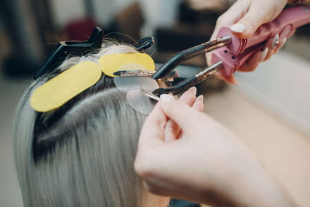 Hairdresser female making hair extensions to young woman with blonde hair in beauty salon. Professional hair extensionの写真素材
