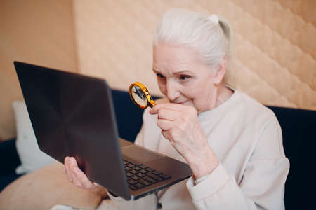 Elderly woman grandmother learns to work at home on laptop computer and internet with magnifier.の写真素材
