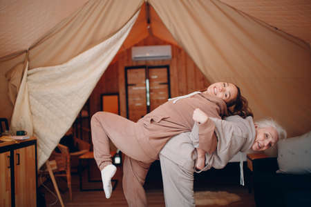 Family doing exercises sports indoors. Young and senior elderly woman relaxing at glamping camping tent. Mother and daughter modern vacation lifestyle conceptの写真素材