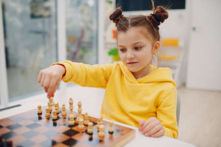 Little girl hold chess figure in hand above chess board. Child playing chess at kindergarten or elementary school.の写真素材