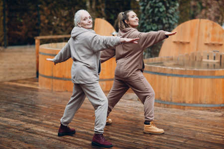 Young and elderly woman warming up and yoga exercises outdoor. Grandmother and granddaughter having zen like modern fitness vacation.の写真素材