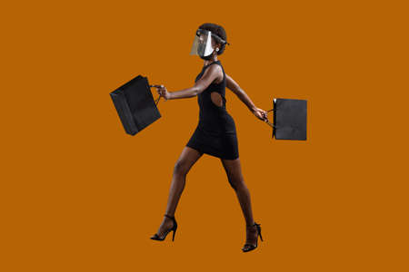 African-american woman full height with afro hairstyle holds black shopping bags and transparent face mask on brown background. Sale and discounts concept.の写真素材