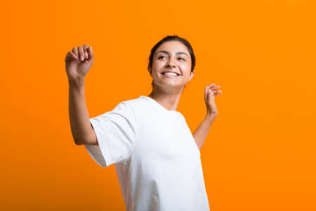 Portrait of young adult happy dancing indian woman in white T-shirt with hands up on orange...