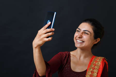 Portrait of positive smiling young adult indian woman in sari having video call with mobile phone and making selfieの写真素材