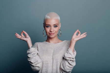 Millenial young woman with short blonde hair portrait sitting and meditate with ok sign mudra on gray blue backgroundの写真素材