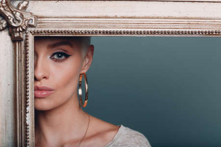 Millenial young woman with short blonde hair on gilded picture frame. Cropped face of beautiful mysterious girl in gold baroque frame corner. Isolated on blue background in studio. Copy space.の写真素材