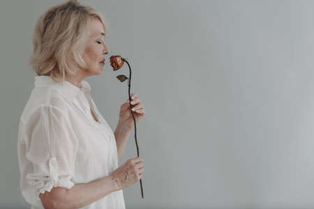 Sad elderly senior woman in white shirt holds withered one dry old dead rose flower. Old age and aging concept.の写真素材