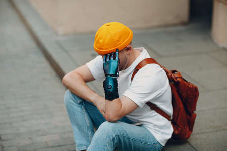 Young disabled upset sad man with artificial prosthetic hand sitting on sidewalk city street outdoorの写真素材