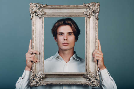Millenial young man artist with blonde hair on gilded picture frame portrait. Isolated on blue background in studio.の写真素材