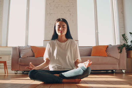 Asian woman doing yoga and zen like meditation lotus pose in casual wear at indoor living room apartment with natural sun light.の写真素材