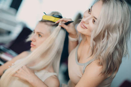 Hairdresser female making hair extensions to young woman with blonde hair in beauty salon. Professional hair extension try checkの写真素材