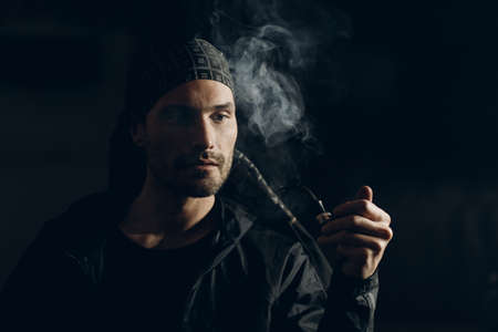 Man smokes smoking pipe against dark background. Back side lit profile portrait with smokeの写真素材