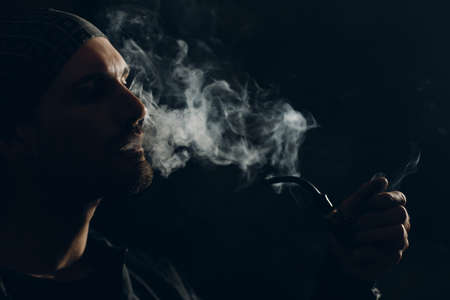 Man smokes smoking pipe against dark background. Back side lit profile portrait with smokeの写真素材