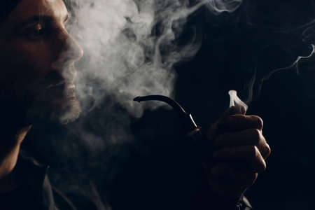 Man smokes smoking pipe against dark background. Back side lit profile portrait with smokeの写真素材