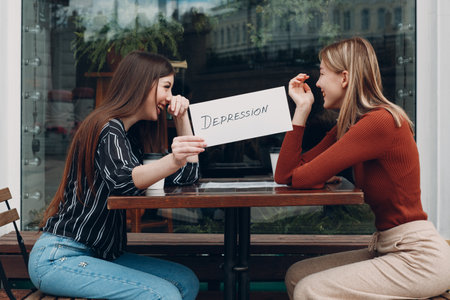 Hidden depression concept. Woman holding white sheet paper labeled word Depression in hand. Two women talking and smiling in street cafeの写真素材