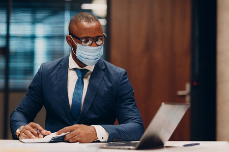 Portrait smiling african american businessman in medical face mask sit at table for meeting in office sign contract agreement with pen and laptopの写真素材