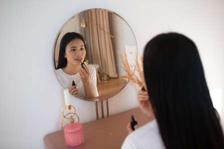 Beautiful Asian woman front of mirror applying make up lips with pink lipstick at living room.の写真素材