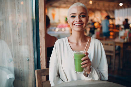 Portrait of young smiling millenial european short haired woman looking through glass with green smoothie at cafe. Beautiful happy blonde girl indoor.の写真素材