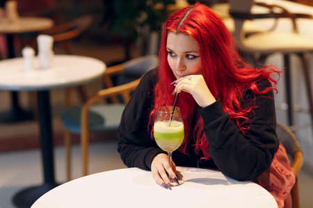 European plus size woman drink green smoothie in cafe. Young red pink haired body positive girl sitting at restaurantの写真素材
