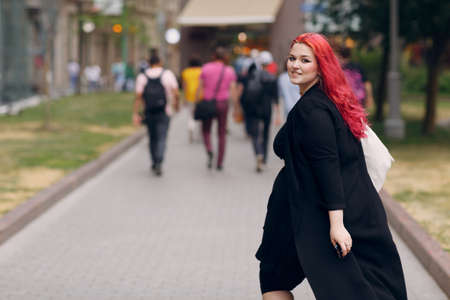 European plus size woman walking outdoor street city. Young red pink haired body positive girl.の写真素材