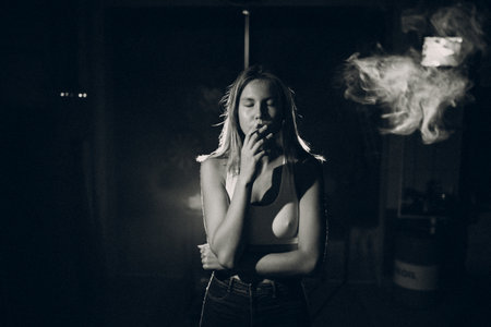 Young woman smokes cigarette in the dark with light spot ray. Black and white image with film grainの写真素材