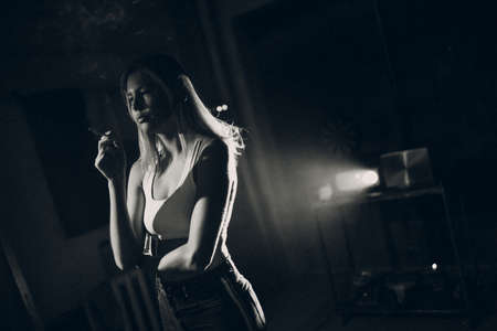 Young woman smokes cigarette in the dark with light spot ray. Black and white image with film grainの写真素材