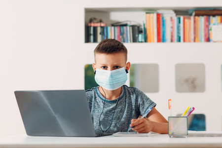 Young boy in headset sitting at table with laptop and medical face mask and preparing to school. Online education concept schoolboyの写真素材