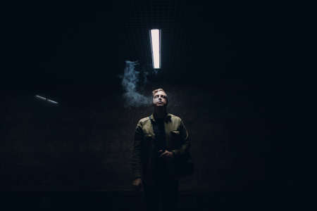 Portrait of young caucasian man smoking e-cigarette in dark with line light.の写真素材