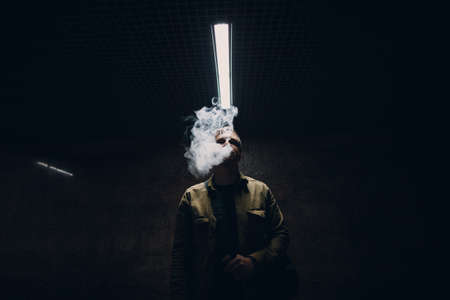 Portrait of young caucasian man smoking e-cigarette in dark with line light.の写真素材