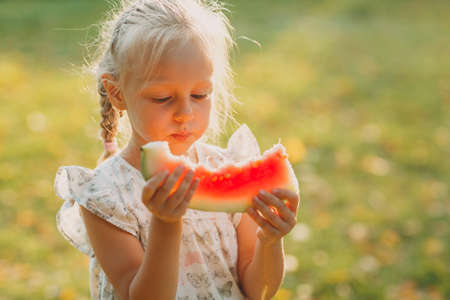 Cute blond little girl with watermelon on the grass in park.の写真素材