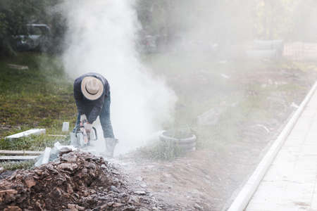 Worker mason cuts the sidewalk tile with circular saw while repairing sidewalk in city street.の写真素材