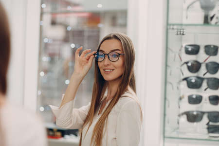Close up of gorgeous young woman smiling picking and choosing glasses at the optician corner at the shopping mall. Happy beautiful woman buying eyewear eyeglasses at the optometrist.の写真素材