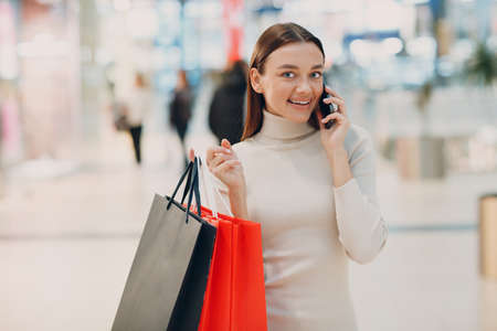 Positive young adult woman carrying paper shopping bags and mobile phoneの写真素材