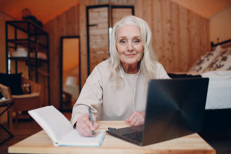 Elderly woman with headphones talking video call on laptop in kitchen, waving at screen, chatting and speaking camera device view.の写真素材