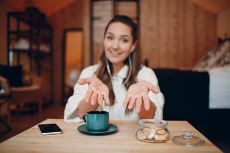 Smiling happy young woman sitting at table at home behind computer laptop and talking on video call. Girl female with cup of tea or coffee speaking online on webcam indoorsの写真素材