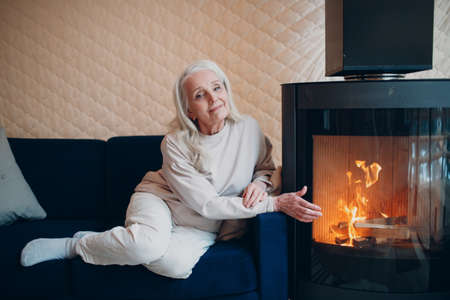 Elderly gray haired woman sitting on sofa in living room with fireplace.の写真素材