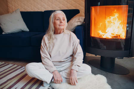 Elderly gray haired woman sitting on sofa in living room with fireplace.の写真素材