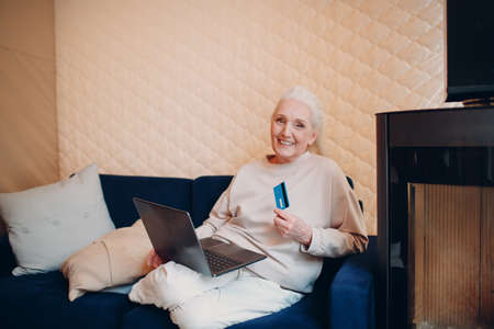 Smiling senior ederly woman using credit bank card and laptop on sofa near fireplace. Online shopping conceptの写真素材